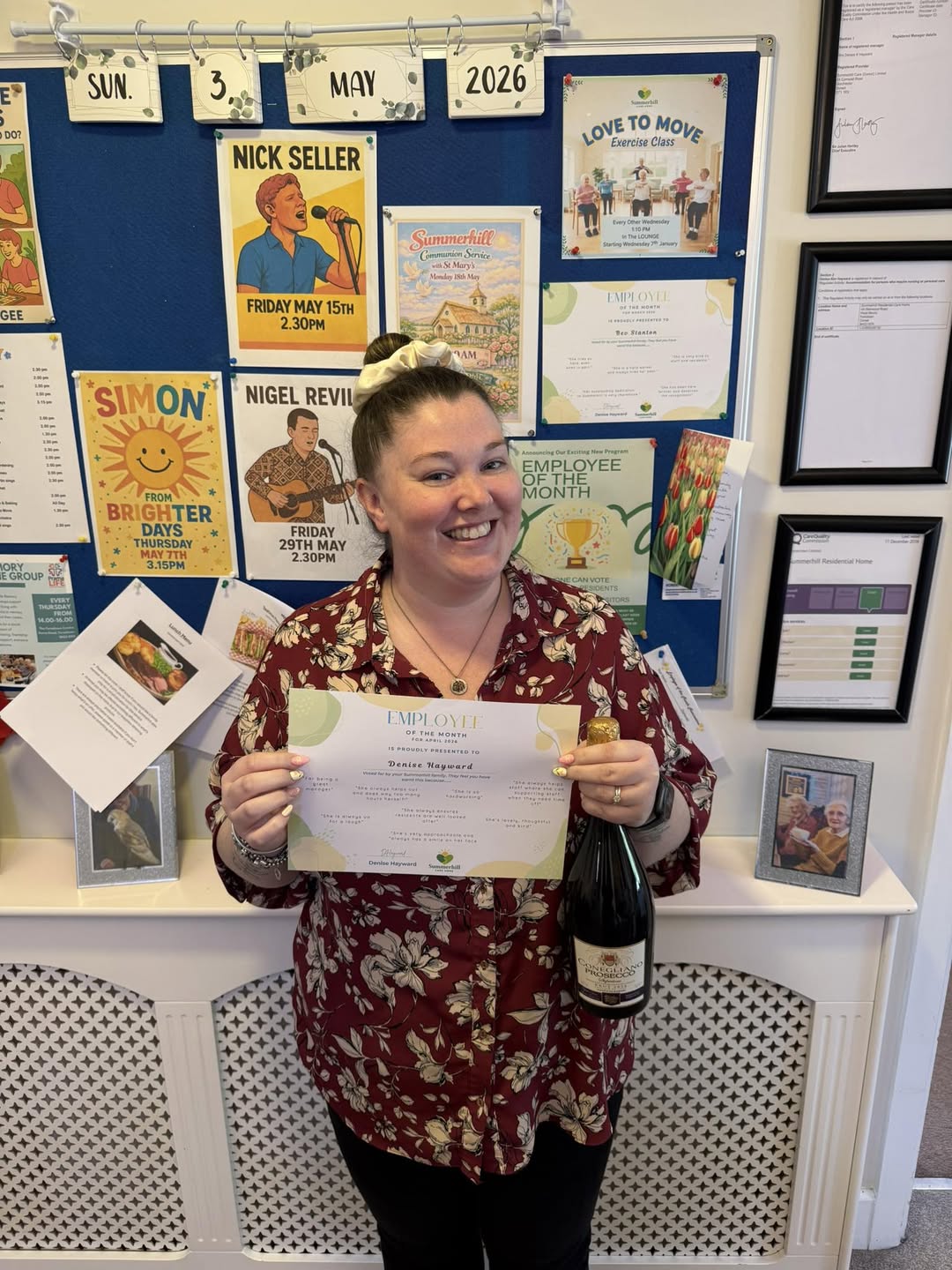 Denise Hayward smiling while holding an Employee of the Month certificate, a bottle and a card in front of the Summerhill noticeboard.