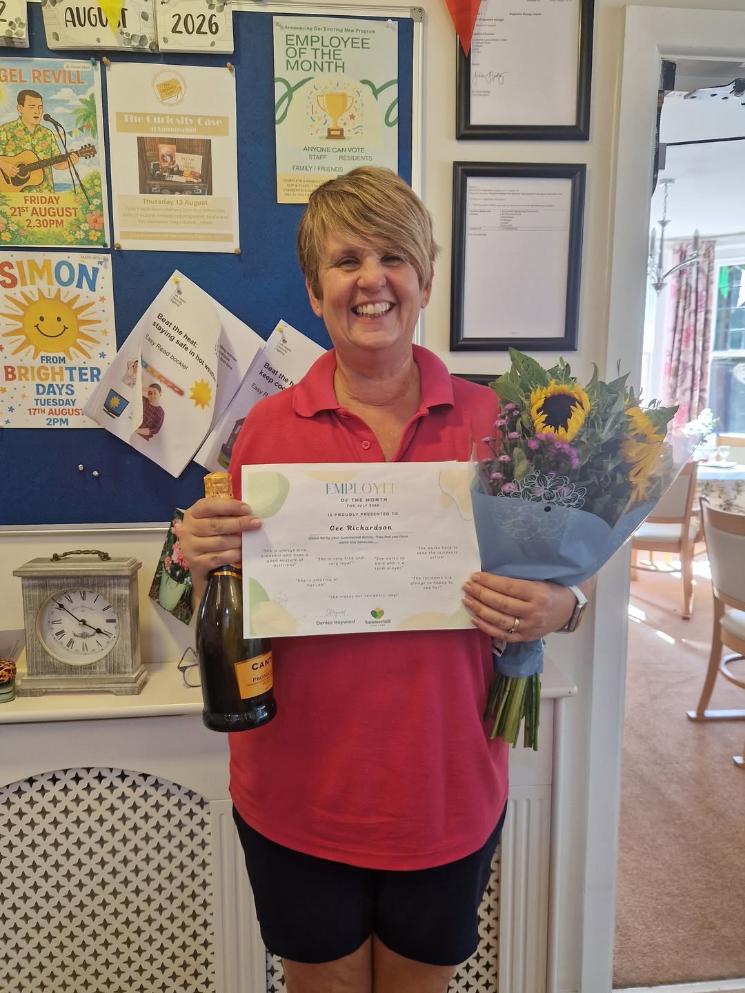Gee Richardson smiling while holding an Employee of the Month certificate, a bottle and a flower bouquet in front of the Summerhill noticeboard.