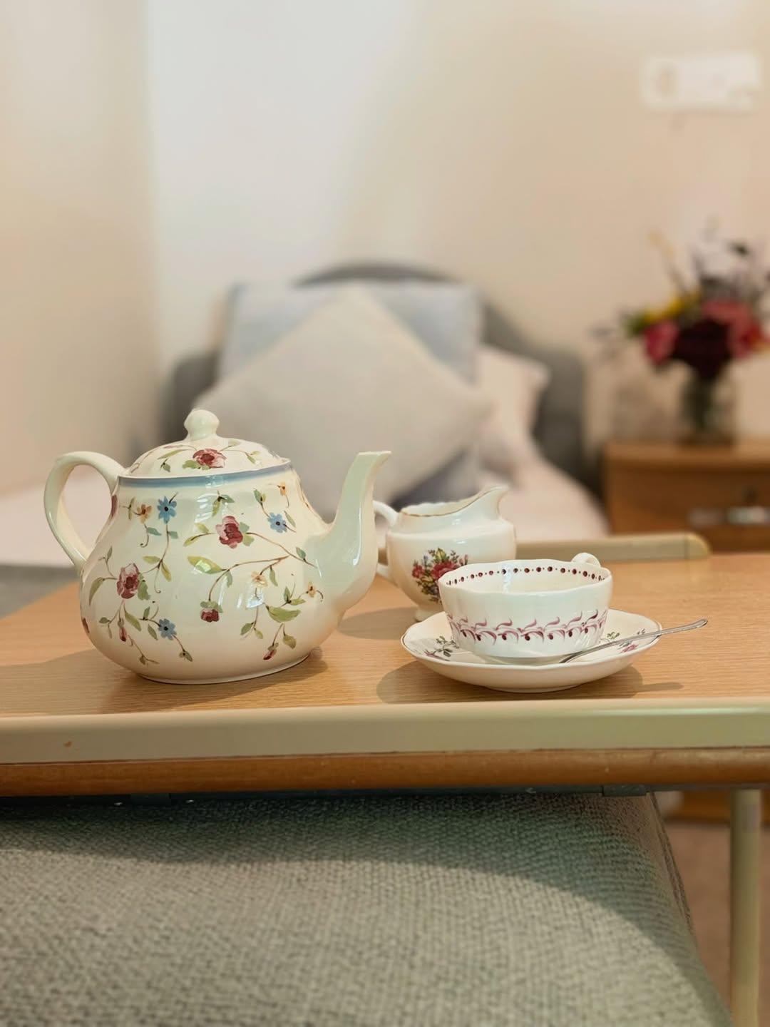 Bedroom detail with a floral teapot, milk jug, teacup and saucer on an over-bed table; the bed and flowers are softly out of focus behind it.