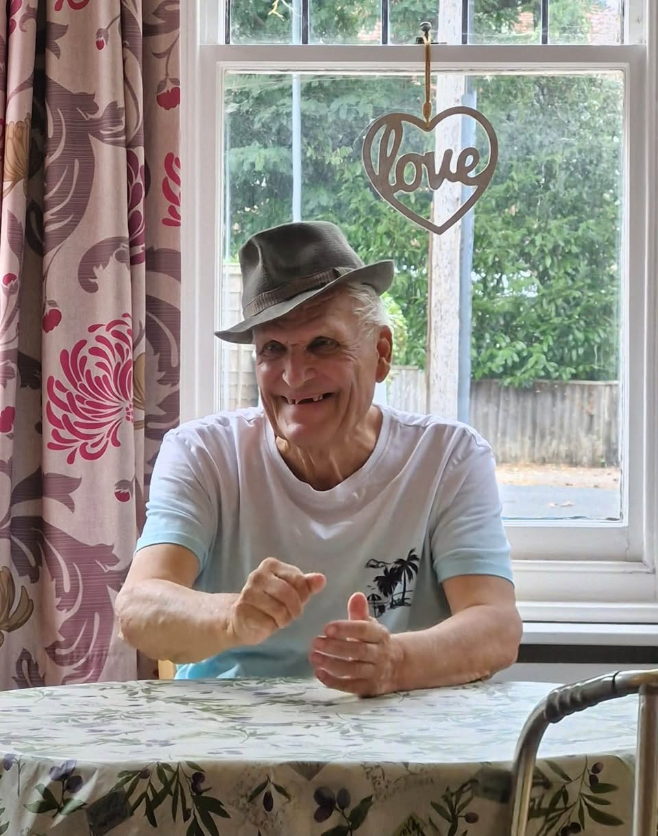 Smiling older man in a fedora seated at a table by a window, with a heart decoration behind him.