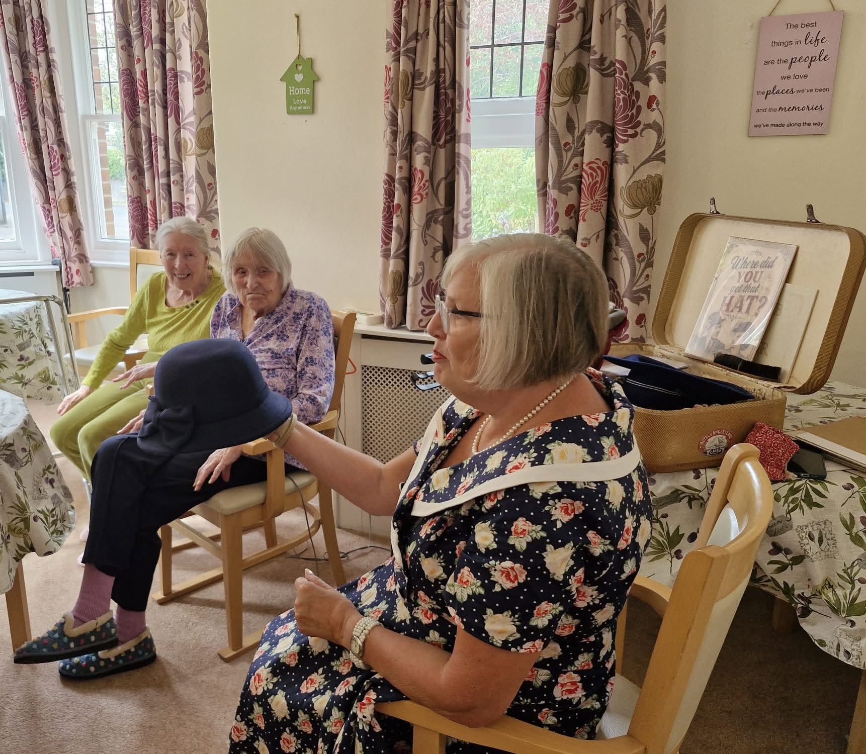 Memory-session scene with a facilitator showing a navy hat to two older women seated in the Summerhill lounge.
