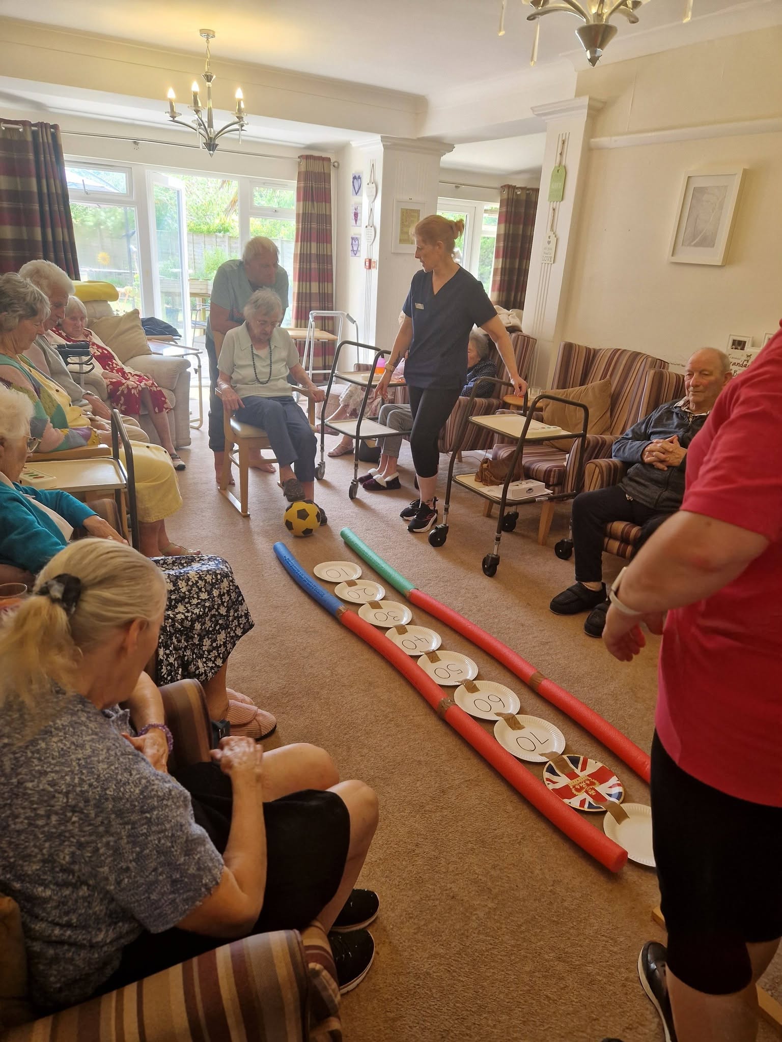 Wide ground-floor lounge view with residents and a staff member gathered around a target-and-ball game, with walkers, seating and garden doors visible.