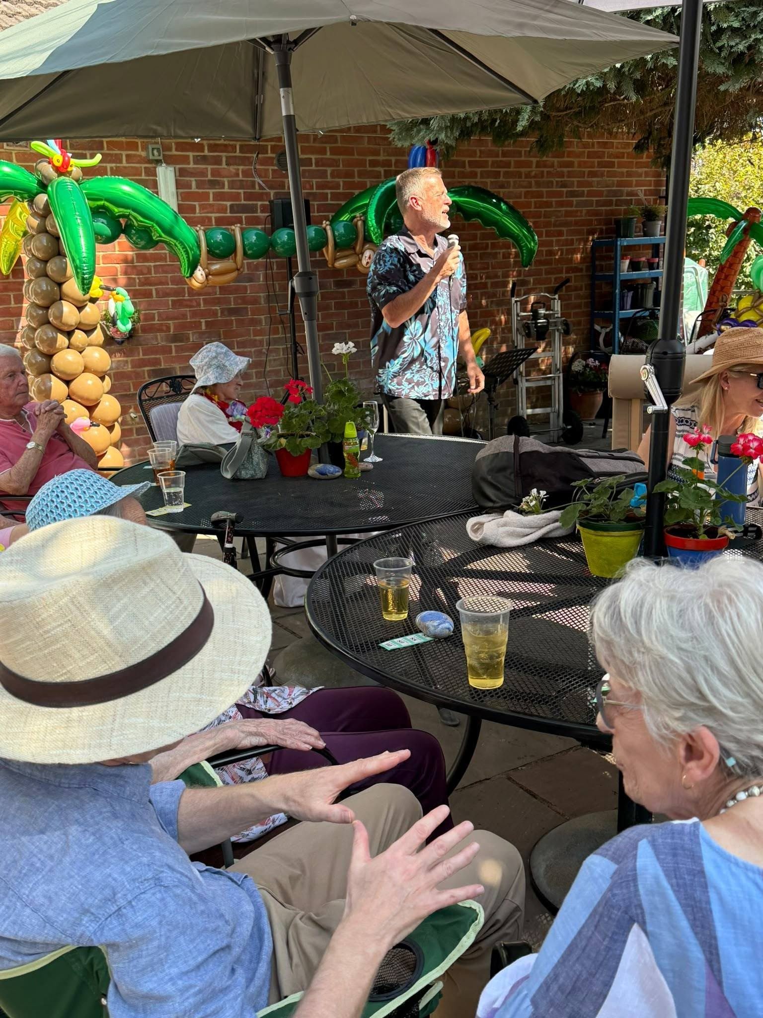 Outdoor Summer Party gathering with a singer holding a microphone beside tropical balloon decorations, tables, drinks and seated residents.