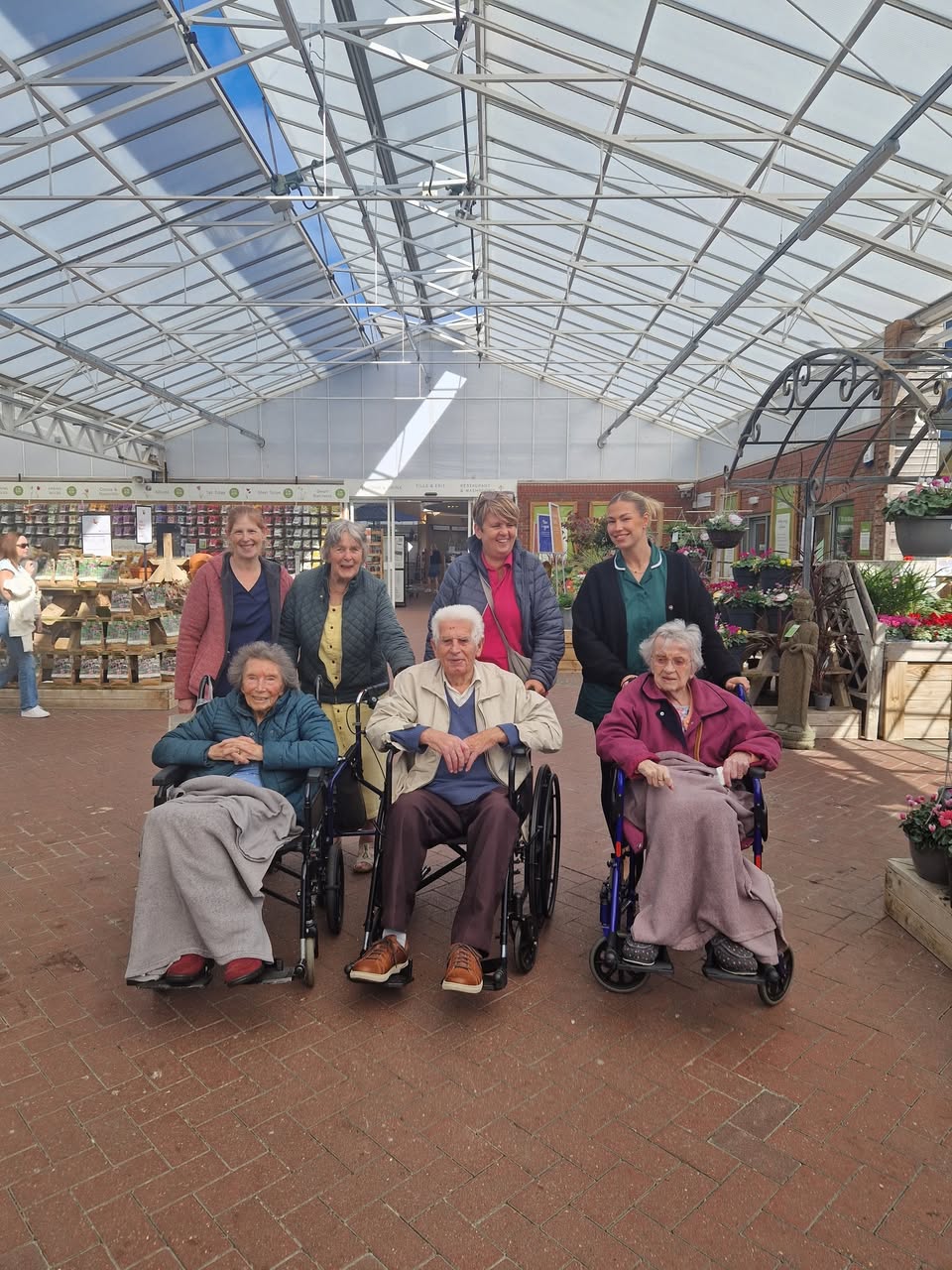 Three Summerhill residents in wheelchairs with four members of the team under the glass roof of a garden centre.
